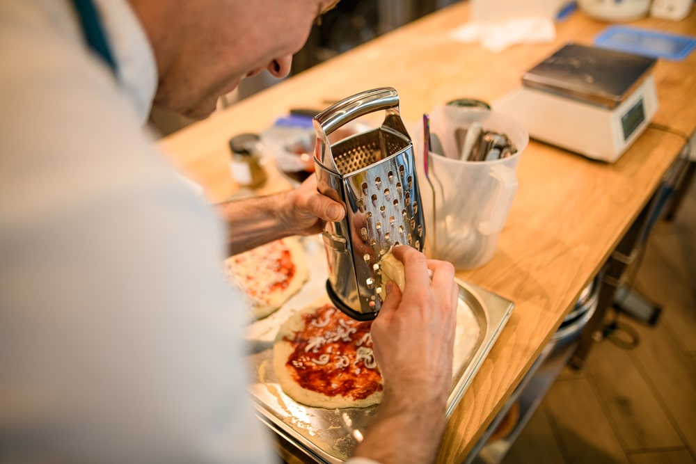 chef holds metal grater in his hand and grates cheddar cheese on pizza